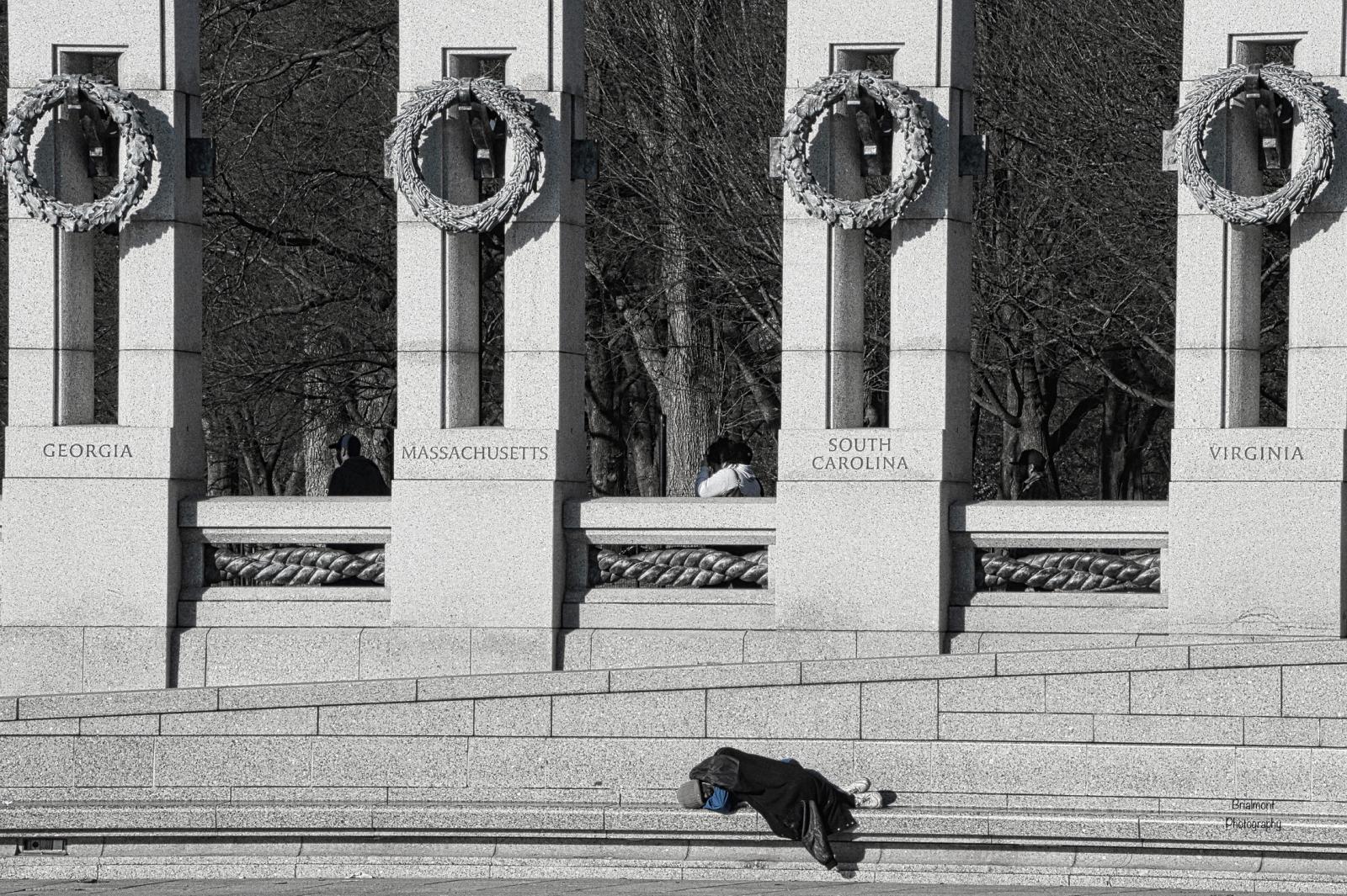 Homeless man sleeping at WWII Memorial while people walk by oblivious.