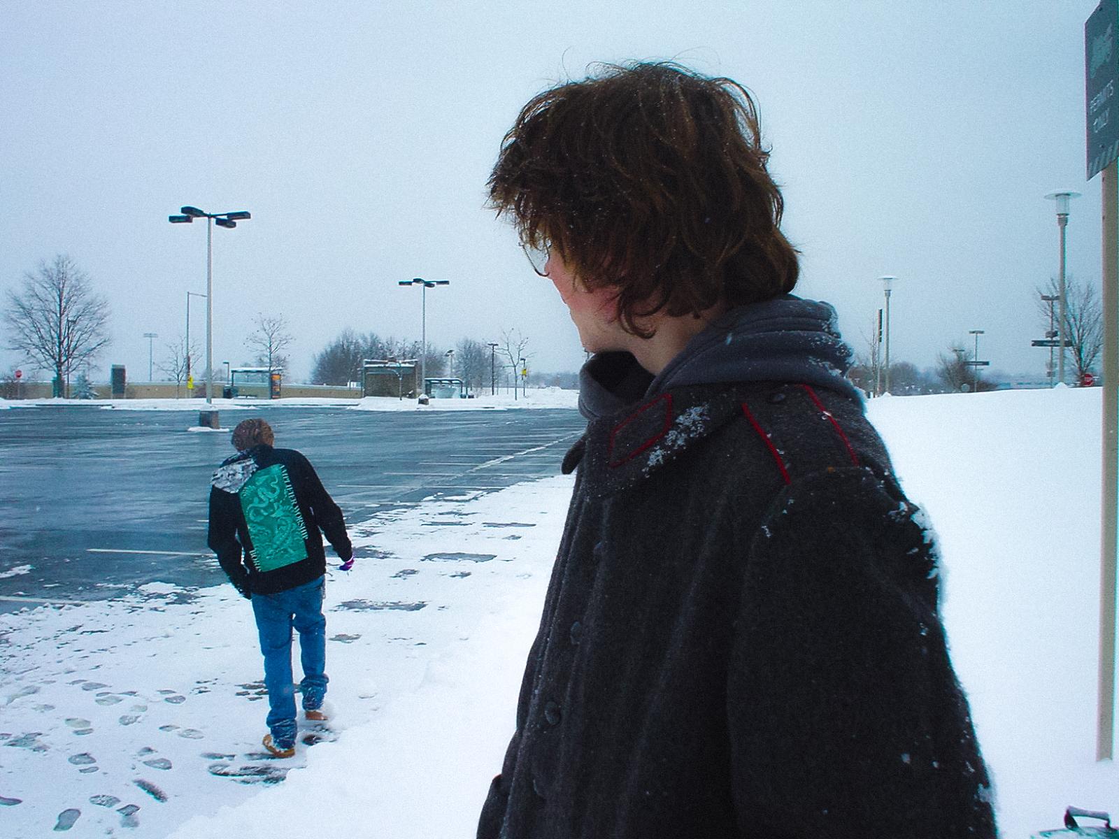 One person walks around in a snowy parking lot while another looks at him