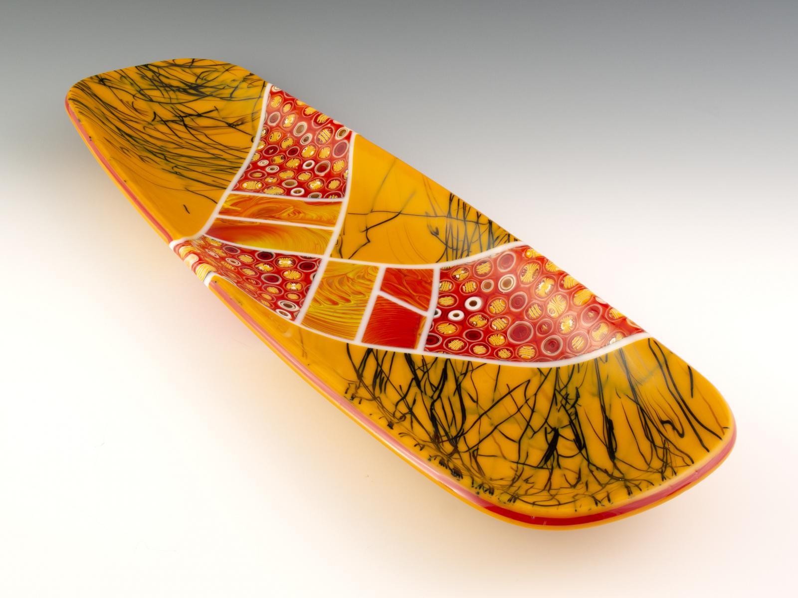 Fat, delicious, juicy cross-sections of hand-pulled patterned glass cane are the star of this bowl, accented with the rippling flow of an aperture pour. The red arrow decisively bisects a plane of black threads in pumpkin-colored glass. The glass cylinders in this bowl are sections individually cut from hand-pulled glass cane, which I create by heating glass to 1500 degrees and pulling it into rods while molten in a modern-day adaptation of the 16th century techniques of the Murano glassmakers.