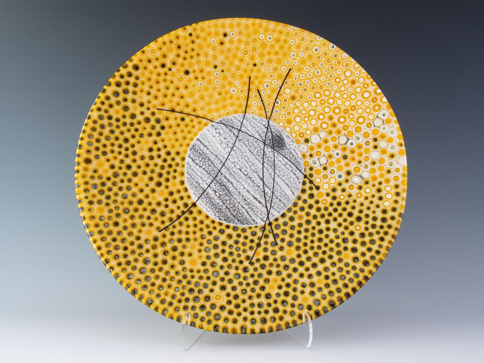 Individual glass threads run through the center of cross-sections of hand-pulled glass cane, with cheerful marigold yellow contrasting with black accents. Across the middle, more curved threads swoop, making a visual connection to the murrine core.
The glass cylinders in this bowl are sections individually cut from hand-pulled glass cane, which I create by heating glass to 1500 degrees and pulling it into rods while molten in a modern-day adaptation of the 16th century techniques of the Murano glassmakers. The gradient of murrine as it moves around the bowl reveals a chronology of the glass cane as it emerged from the crucible.
Fused, cold worked, and slumped.