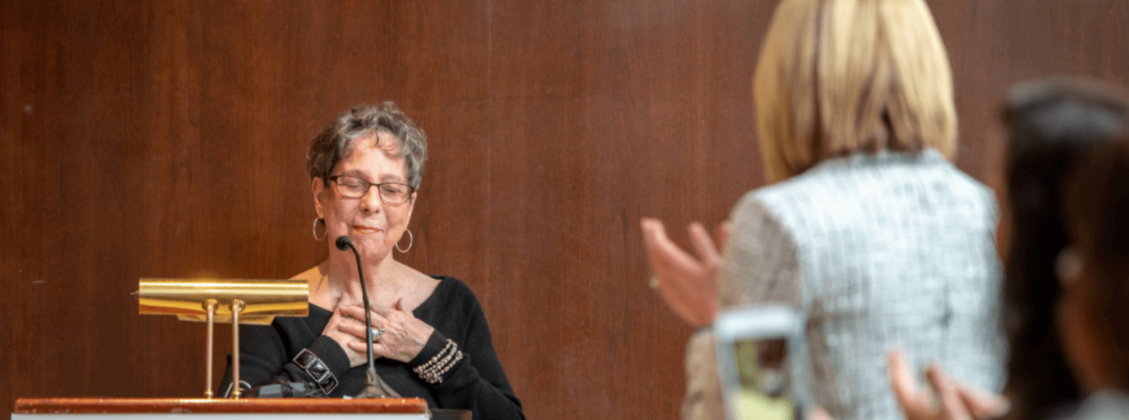 Two people clapping for the Poet Laureate who is standing behind a podium