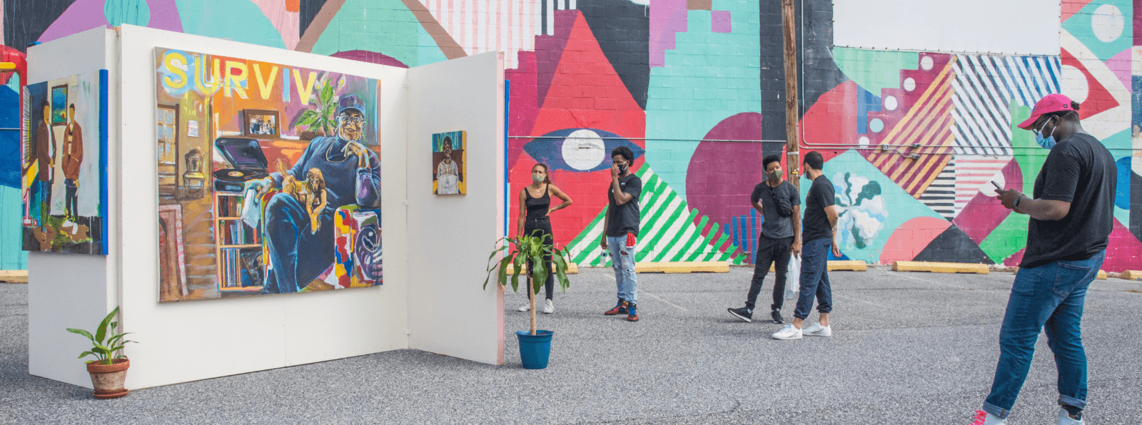 Five people in front of a mural and in a parking lot looking at paintings installed on a free standing white wall