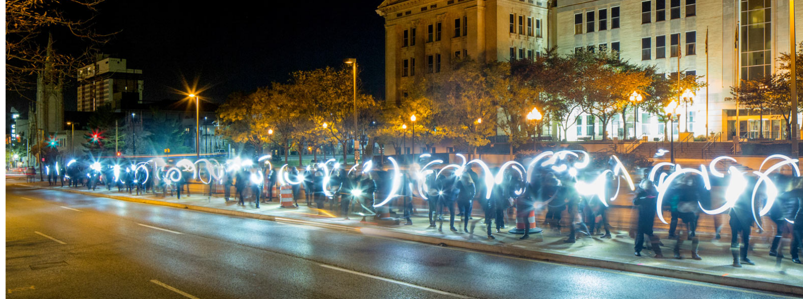A crowd of people on the sidewalk spinning lights in front of a building.