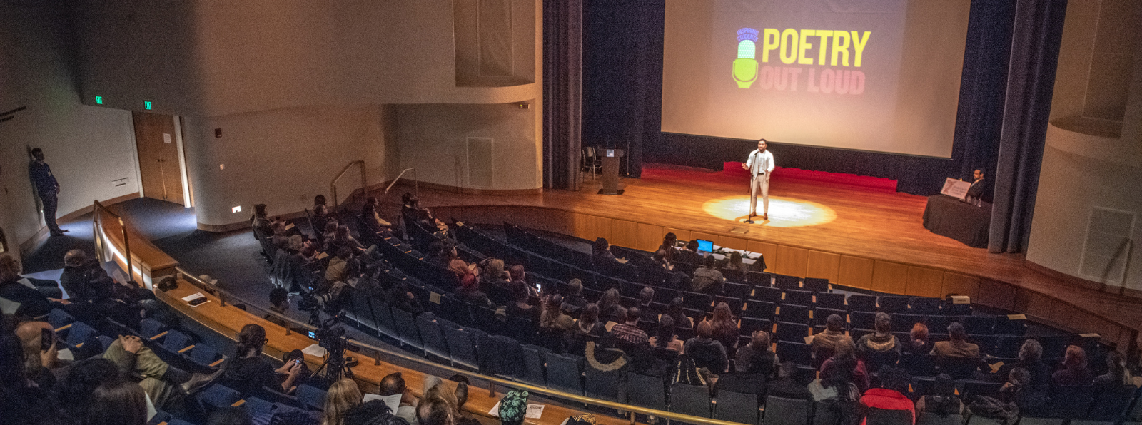 An auditorium full of people are watching a person on stage. “Poetry Out Loud" is projected behind the stage.