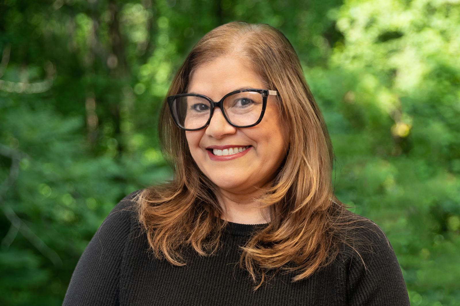 Headshot of Councilor Albita Rivera. Woman with glasses and a black shirt smiling in front of green forest background
