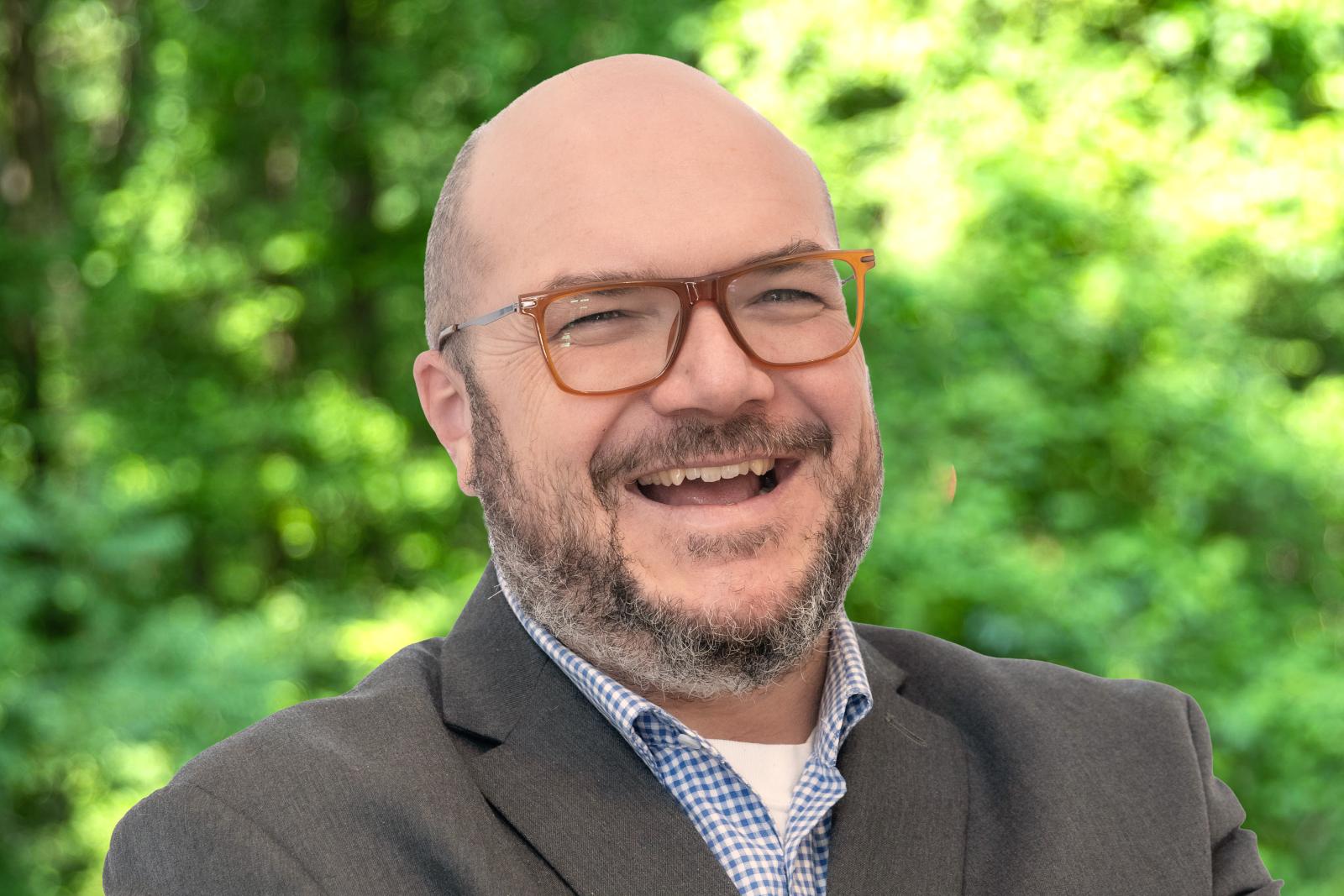 Ryan Patterson headshot. Smiling man with glasses, a mustache and beard, and wearing a grey sport coat over a light blue shirt.