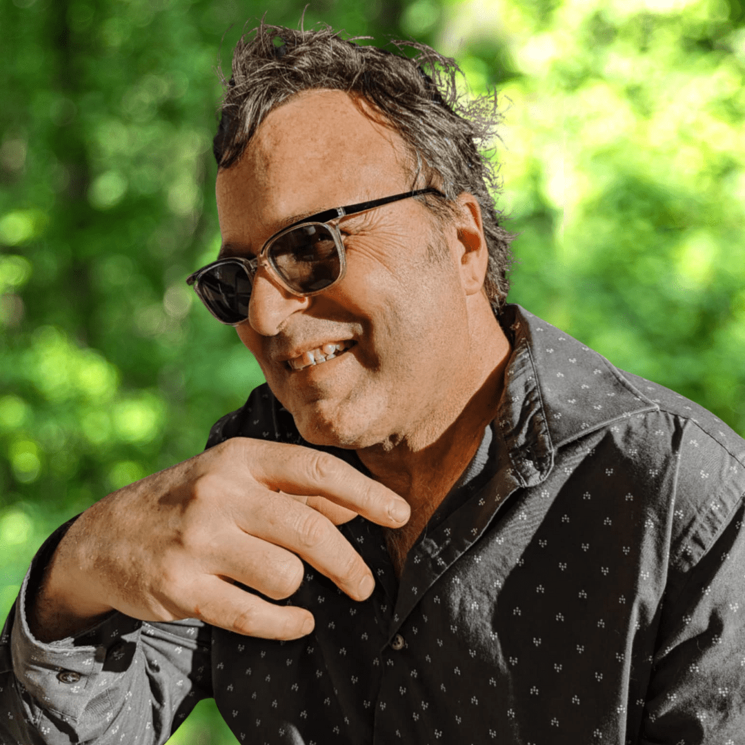 Smiling man with wavy salt-and-pepper hair and dark glasses, wearing grey shirt with white pattern