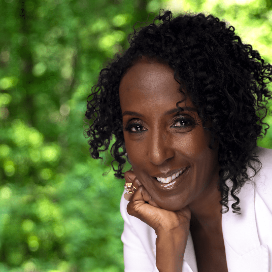 Smiling woman with curly black hair resting chin on closed hand, wearing a white suit jacket and looking directly at the camera.