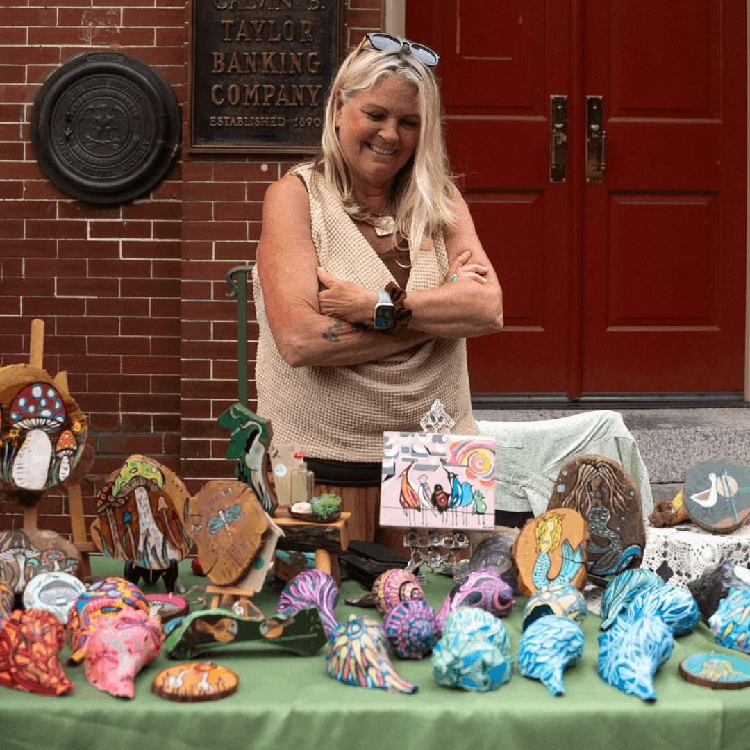 Women in tan sleeveless top with arms crossed smiling at vendor's table full of artwork