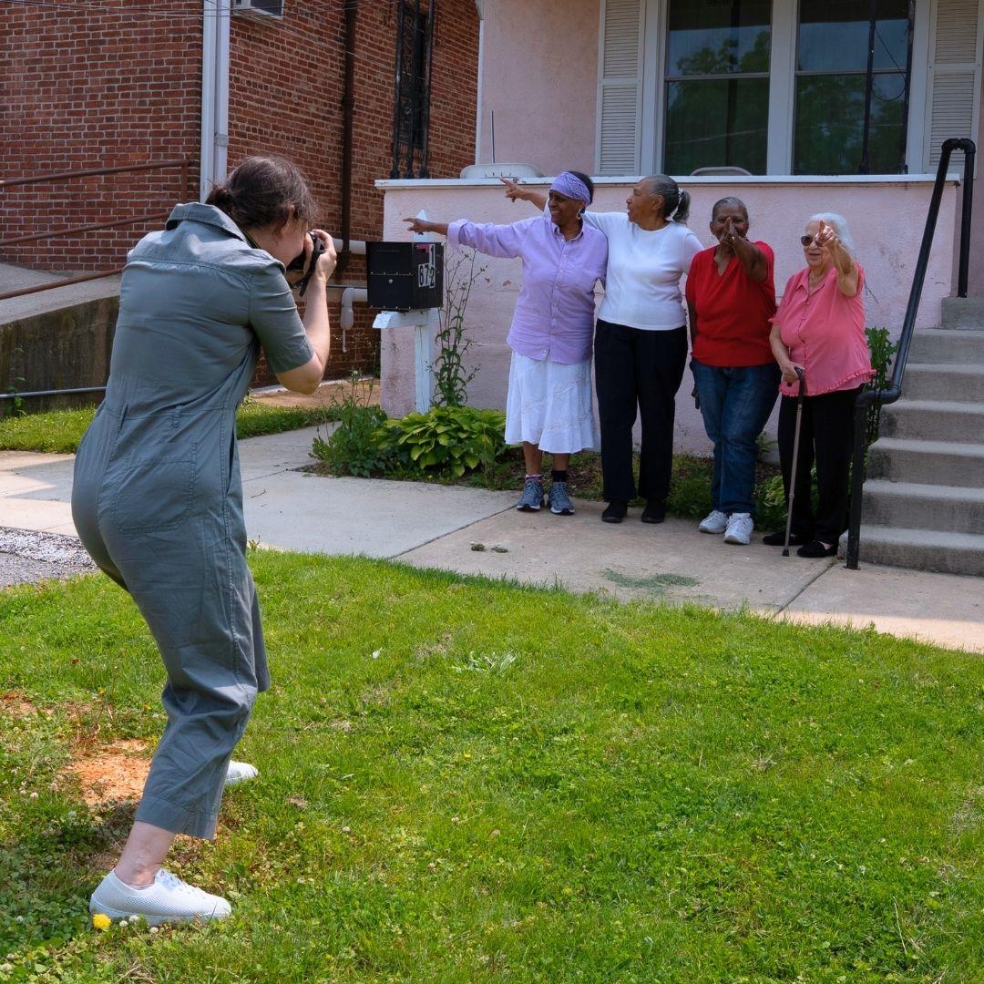 Eva Marquez Salmeron in a gray jumpsuit photographs four older women standing and pointing their index fingers in different directions on a sidewalk outside the A.M.E. Church Outreach Development Center.