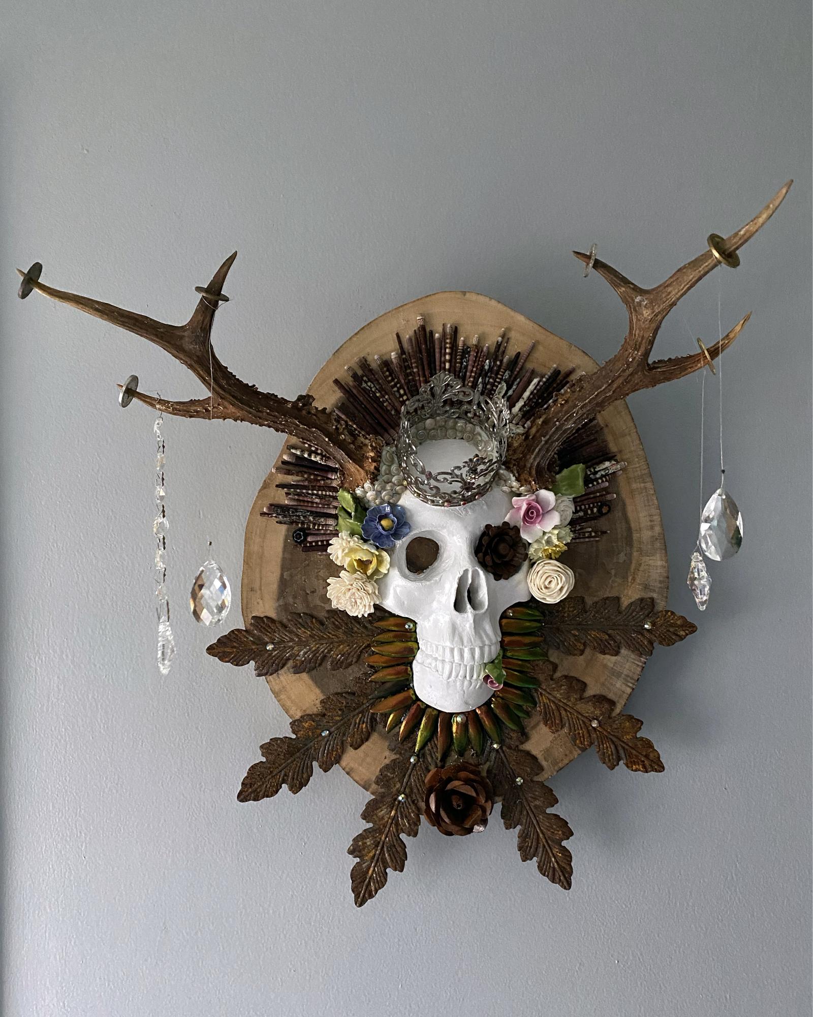 plaster skull with crown on acacia wood with rusted leaves and flowers, roe antlers, beetle wings, urchin quills, ceramic and wood flowers, crystals, and rhinestones