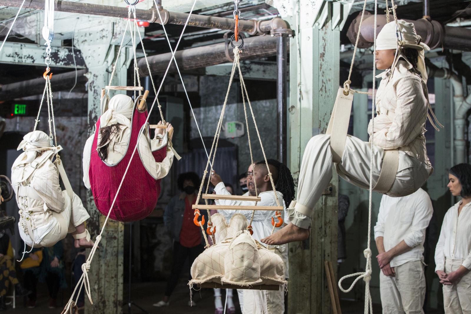 Four people, including myself are suspended in varying positions from supine to upright in harnesses and slings using geared rope-pulleys counterweighted by sewing machines, they are in turn tended by four other people.

I use a dozen sewing machines ranging in size and mass from 150lb (7-class harness machine) to around 50lb (31-class standard early 20th cent. Industrial machine) to 20lb early home sewing machine to the singer featherweight portable machines which weight under ten pounds, while they differ in size they share the essential “Black Singer” form and nostalgically remind us of everything from domesticity and polite accomplishment (with all of its complex problems of unpaid work and patriarchy) to industrial sweat-shop labour.

The industrialists in the heat of the Industrial Revolution understood that due to technological advancement, their machines would be redundant in as little as three years. In order to justify their investment, they would have to run their machines day and night, which led to 24-hour shift work (which is in itself inhumane), the labour shortage that ensued, precipitated an explosion in child labour in Europe as well as fostering the dehumanizing notion that people are inferior to capital.

A notable illustration of this devaluation of human life was the 1911 fire at the Triangle Shirtwaist Factory, New York City’s deadliest industrial disaster claiming the lives of 146 garment workers, mostly recent immigrants between ages 16-23, many of whom jumped to their deaths because the exits were locked to prevent theft and unauthorized breaks. 

In Bangladesh in 2013, Rana Plaza collapsed, killing 1134 garment workers. The building had been declared unsafe. Banks and stores in the plaza were closed, but the 5000 garment workers were threatened with termination if the did not show up for work. 

As consumers, we are at least culpable in this continuing tragedy
