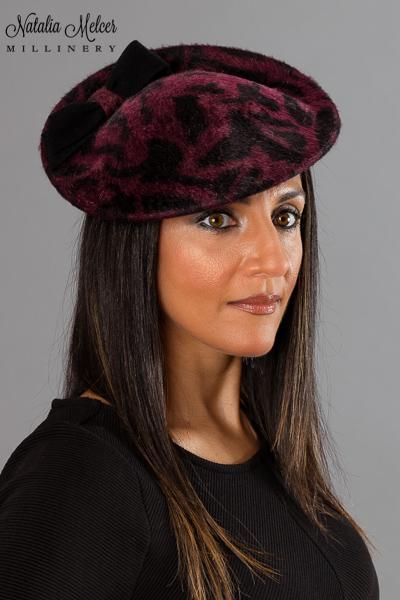 Maroon jaguar print fur felt perching hat adorned with a black fur felt bow. Hat was made by blocking the felt over a wooden form and the hat is secured with an elastic band that goes under the hair. One-of-a-kind, handmade hat using traditional millinery techniques.