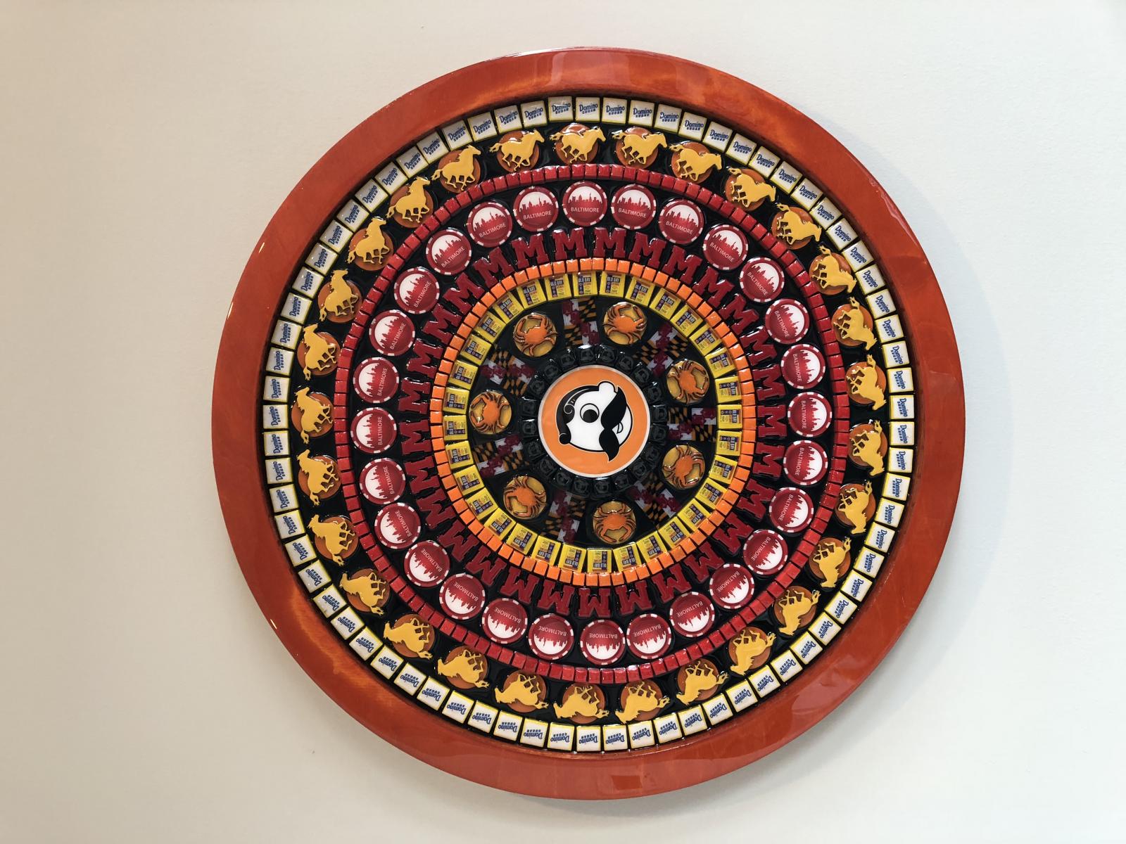 This 36 inch diameter nostalgic collage is titled, “Be-More.’ It is composed of hundreds of items that are iconic from Charm City Baltimore. The center eye serves as the anchor of the piece and it is the iconic “Natty Bo” symbol from National Bohemian beer. It is surrounded by Maryland flags and crabs. In Baltimore, having beer and crabs, seasoned with Old Bay from the McCormick Spice Company, is a long-held tradition. Pieces included in this work represent regional themes such as horse racing, the flagship university of the state, the University of Maryland, and the iconic Domino Sugars plant, from the heart of Locust Point, Baltimore. I hunt for unique pieces from the past and assemble them into complex collages of texture and color creating “Nostalgic Art.” Although many pieces are vintage, others I create by hand at the Baltimore Foundery. Each piece is positioned strategically to create a pattern that draws the work together. Planning takes several months for a new design. Assembly begins in the center and I work outward with construction taking place over several weeks. The completed pattern is then sealed using multi-layer epoxy resin, which creates shimmer and protects the work. The resin also allows you to trace your fingers over the different textures, for both a visual and tactile experience.