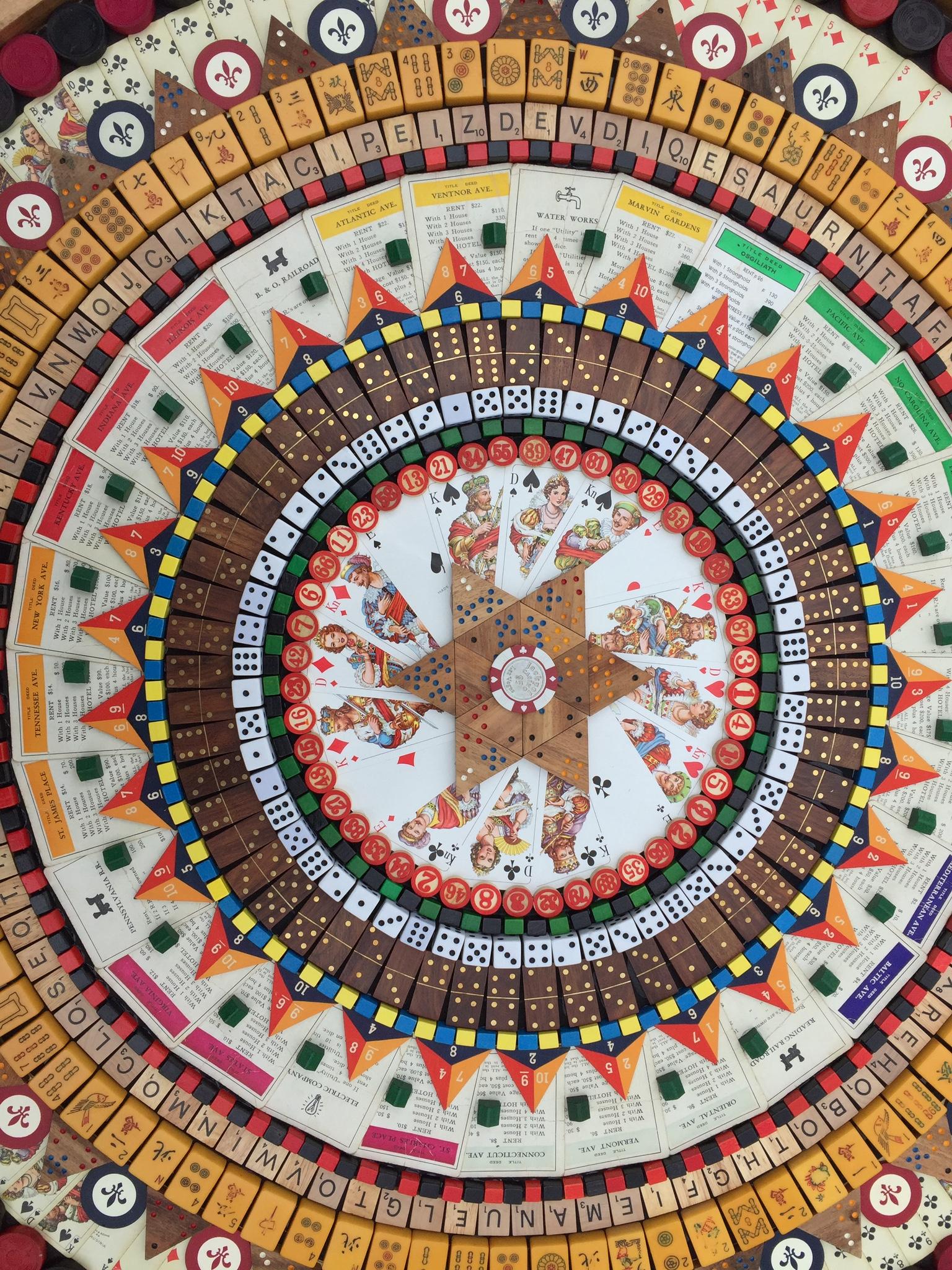 Vintage game pieces to include monopoly, checkers, mah jong, scrabble, and more arranged in concentric circles on a wood base.