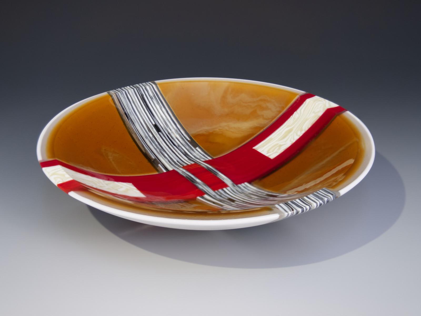 Evoking the glory of autumn, bold swaths of red and vanilla-bronze glass interlock at the heart of this bowl, surrounded by a sea of amber with delicate cream swirls. Windows of clear interspersed within the vanilla-bronze stripes allow light to shine through despite the heft and solidity.

Fused, cold worked, and slumped.