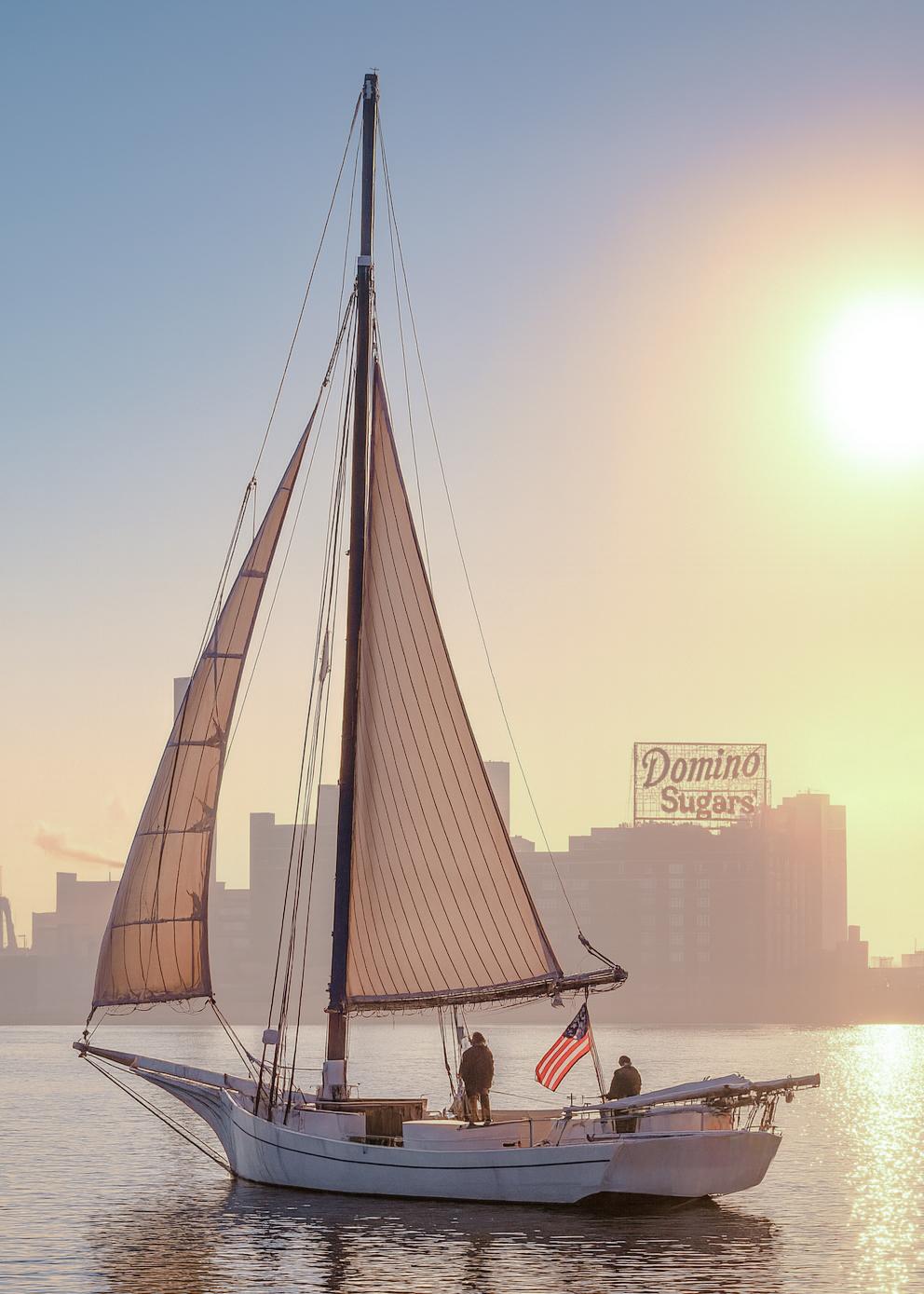 A classic Chesapeake Bay skipjack glides across calm harbor waters at sunrise, its tanbark sails glowing in the golden light. The iconic Domino Sugars sign rises through the morning haze, framing a serene scene that blends maritime heritage with Baltimore’s industrial skyline.