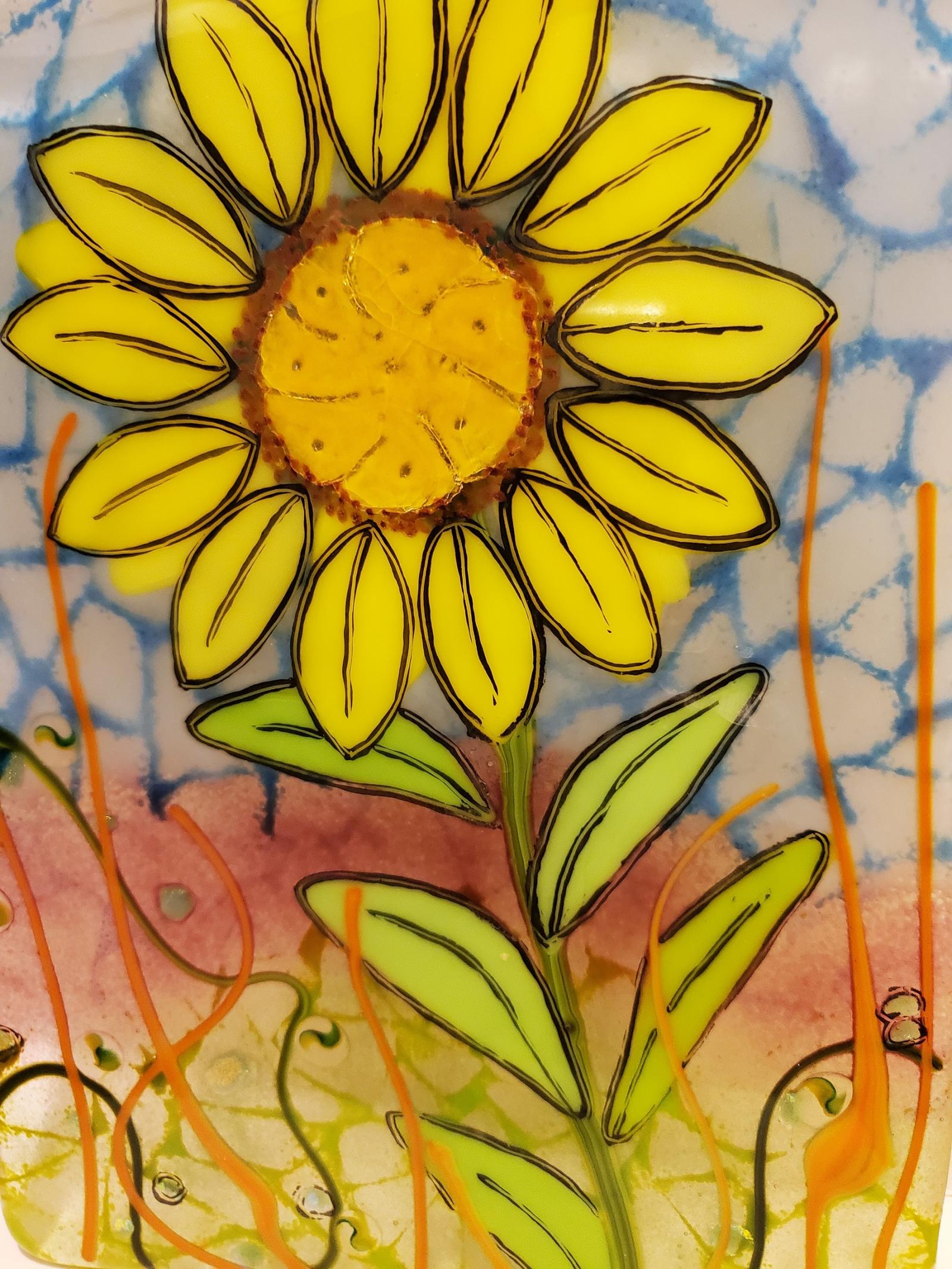 The blind Helen Keller once said, "Keep your face to the sunshine." And, that's just what these radiant sun flowers do - follow the sun.  Encourage in the warmth and feel of sunshine into your home simply by hanging this bright and cheerful fused glass sunflower on your wall. The piece is comprised of fused glass with hand-painted metallic glass elements to make the flower and leaves pop. The piece hangs by a metal wall mount, and is 7" x 9". 