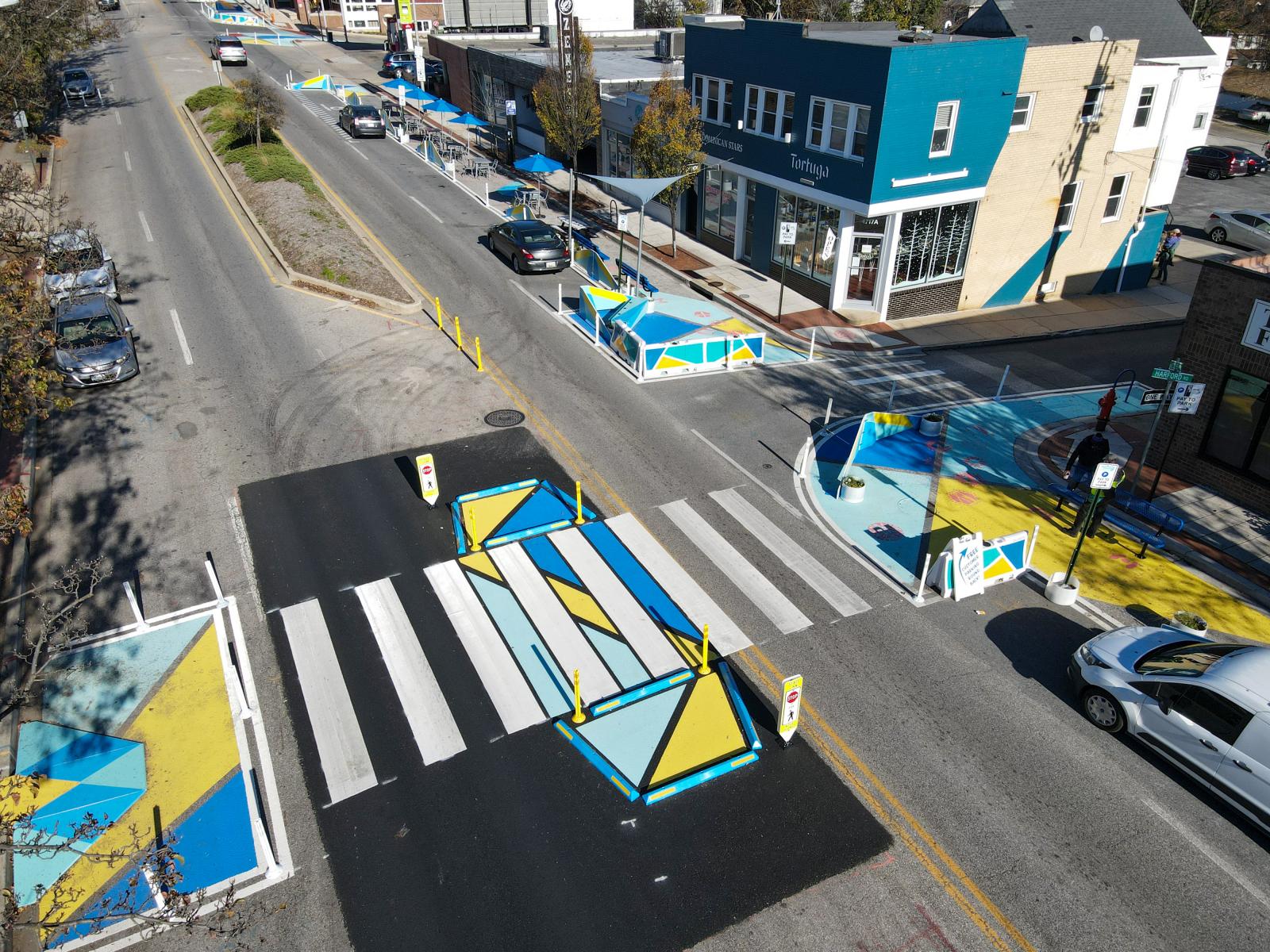 Design for Distancing Curbside Commons
https://vimeo.com/528394639
https://grahamprojects.com/projects/curbside-commons/ 
StreetBond 150 pavement coating, thermoplastic crosswalks, and bump-outs, flex-posts, water-filled barriers, shade sails, outdoor seating, umbrellas, custom benches, bike racks
15,400sf, dimensions vary
June - November 2020
4300, 4700, & 4800 blocks of Harford Rd, Baltimore, MD 21214
In response to the COVID-19 pandemic, the Curbside Commons Design for Distancing project converted a parking lane into public space for safe, physically distanced community gathering, shopping, services, and culinary encounters along Hamilton-Lauraville’s main street, Harford Road. Led by Graham Projects, the design-build team included Property Consulting, Inc., LANNINGSMITH, and Annie Howe Paper Cuts. The team collaborated with the Hamilton-Lauraville Main Street and adjacent small businesses to create public spaces that meet their needs to stay open while maintaining COVID-19 precautions, including outdoor seating, distancing markers, event space, pedestrian and wheelchair accessibility, public art, signage, bicycle parking, and artful wayfinding.
Project partners: Hamilton-Lauraville Main Street, Baltimore Development Corporation, Neighborhood Design Center, Baltimore City DOT, Equus Striping