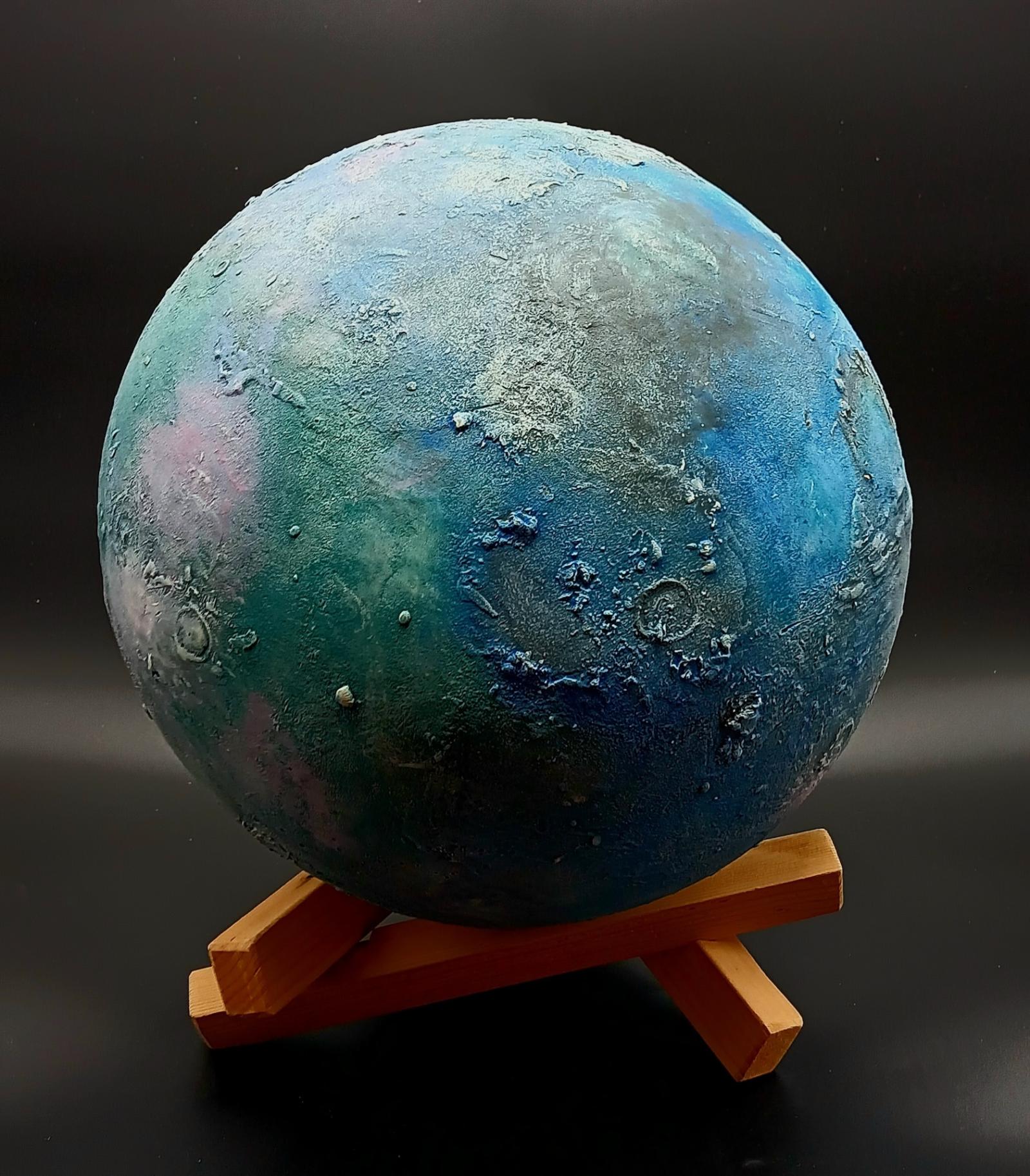 A 14 inch diameter ceramic sphere on a wooden tripod. The piece is sculpted in detail to look like an imaginary moon that just might be out there someplace once the Webb telescope finds it. But for now, imaginary will do. A layered treatment of the surface simulates craters, ice flows, escarpments and small debris, while pigments (underglazes) are layered to represent different mineral deposits on the surface. Ideal centerpiece for a home or office, with special lighting to show off the color. Very nice piece, very finely crafted.