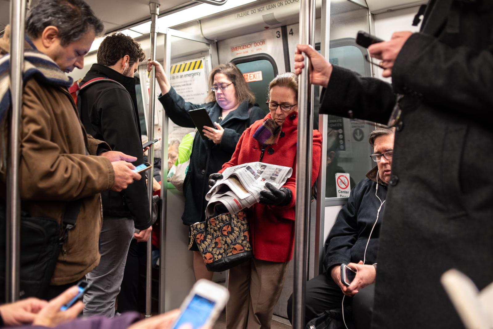 In this photograph, a standing woman in a red coat on the Metro underground reads a newspaper surrounded by eight people reading on electronic devices.