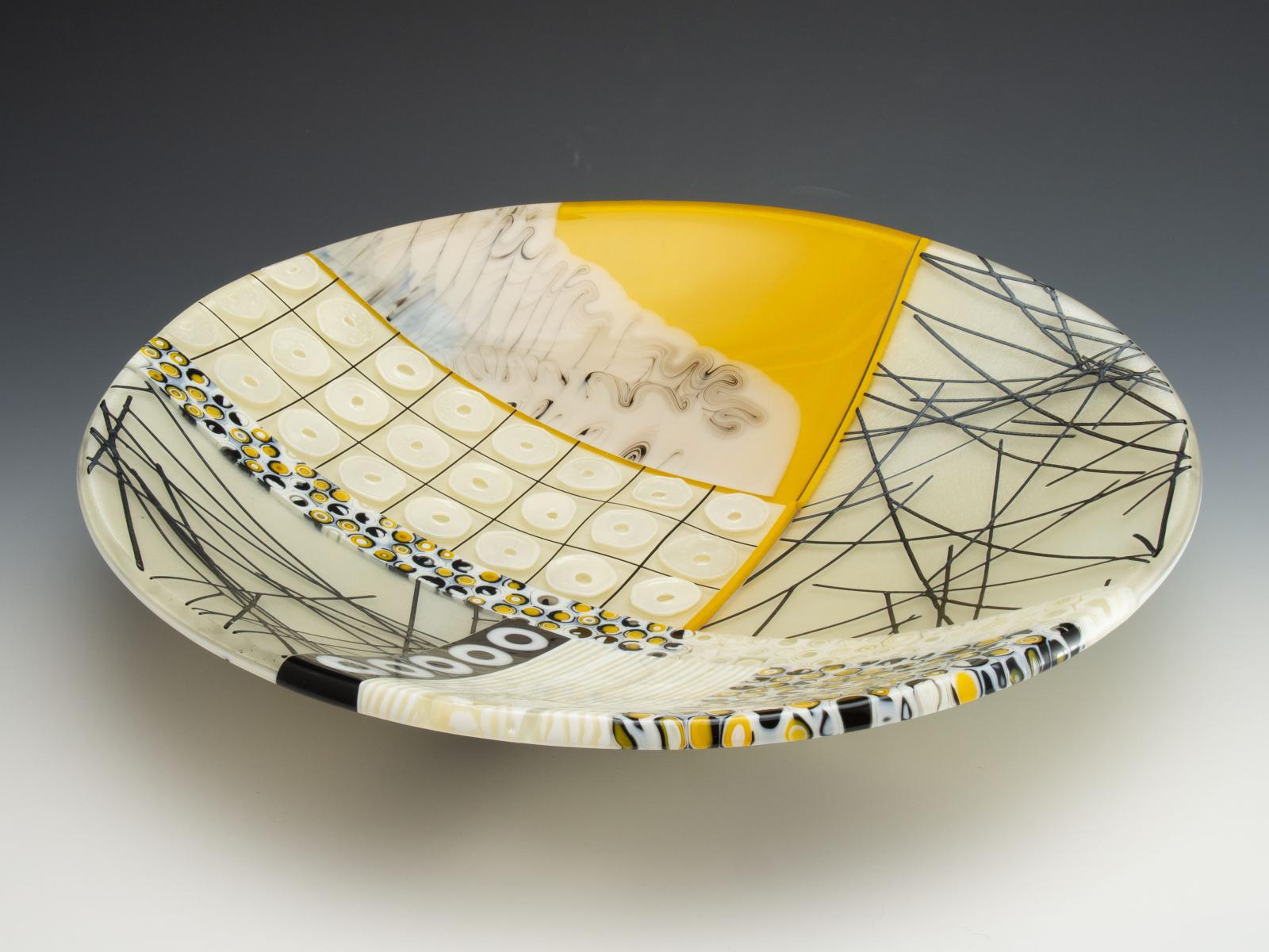 Taffy-like ripples of black and white over marigold yellow, complex patterned murrine, floating threads of glass, and raised rings of glass powder. You'll want to run your fingers over them every time you walk by.

Threads of black glass, scattered over creamy vanilla, one segment shining with iridescence, set the tempo for the symphony. Look into the edge, and you can look under the black glass threads!

You can't help but touch the three-dimensional wafers of powdered glass, marching in time over a grid of floating lines.

Segments of patterned glass cane sport swirling half-circles and juicy yellow centers. Others contain complex patterns of creamy lines in their centers. Along the rim, you can look into the middle of the cane.

In one quadrant, taffy-like ripples of black and white float over marigold yellow. Across the bowl, stripes of parchmenty vanilla surround a floating block of melted and rippling cream.

On the back, strands of black glass are scattered. There's something new to see every time you look at the composition.

The glass cylinders in this bowl are sections individually cut from hand-pulled glass cane, which I create by heating glass to 1500 degrees and pulling it into rods while molten in a modern-day adaptation of the 16th century techniques of the Murano glassmakers.

Fused, coldworked, and slumped.