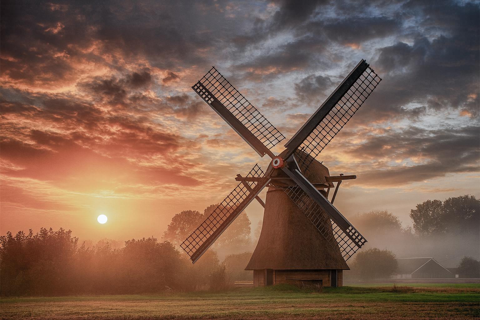 A solitary windmill stands in quiet majesty as dawn breaks, its dark silhouette etched against a sky ablaze with gold and crimson. Mist drifts across the fields, softening the edges of light and shadow. The scene evokes timeless grace — a harmony between human craft and the serene rhythm of nature.