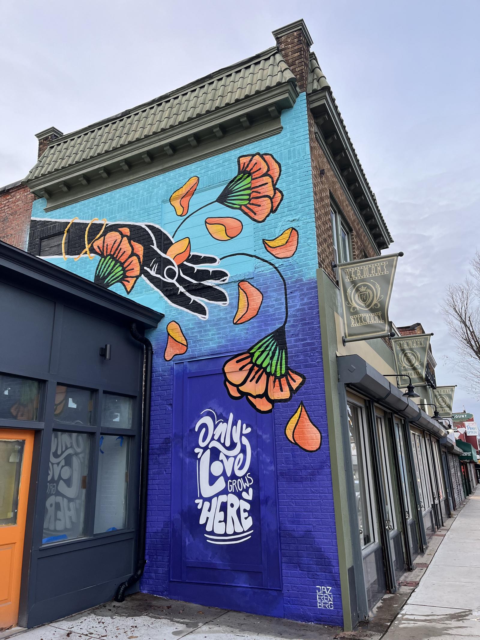 This dreamy mural depicts the act of ‘growing love’. A large black hand tosses orange marigold petals that fall into the night sky, igniting the viewers’ imagination, and transporting them to a world built on love. Huge bright orange marigolds symbolize love and passion, the petals sprinkled into the starry night sky, like releasing a wish or planting a seed.
Viewers are invited to stand on either side of the text “Only love grows here”, which is positioned to fit into the framed panel that is already on the wall.
Location: 3106 Greenmount Ave. Baltimore, MD