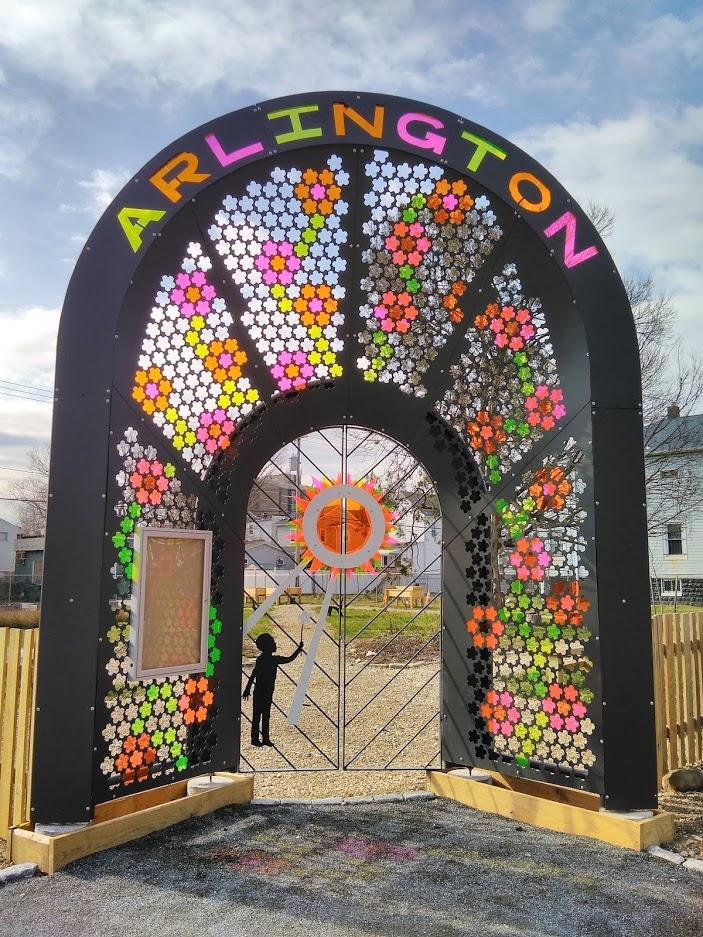 Created as an entryway for Arlington Elementary’s INSPIRE garden park, this piece is an altar to the Sun which provides us with all we need to grow and learn.
Throughout this project FSC worked with community leaders to make a gate that would show the community that they too deserved something beautiful to enjoy.
A steel arch bends 15′ over head. In the center a child offers a flower as a thank you to The Sun.
Resembling stained glass, the light from the sun casts colorful shadows on the ground through acrylic flowers. The mesh panels feature hundreds of 5 sided plasma cut flowers. Originally cut to hold the ends of reclaimed 2L soda bottles, we adapted the design after speaking with community leaders to create something even more magical.
This was one of several major projects FSC took on in 2019. To pull off these projects we purchased a Sprinter van and brought on Thabo Letsebe to assist during the busy September-October crunch. In the end, I decided to title the piece Thank You For Helping Me Grow.
