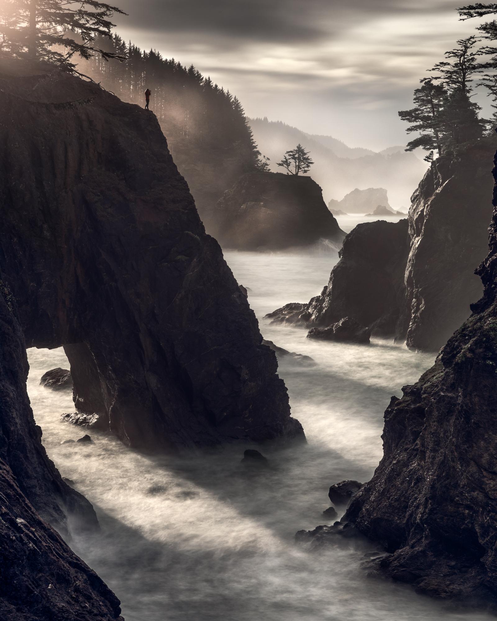 I love light. And getting the opportunity to chase light along the Oregon Coast is just straight-up magical. However, sometimes it never shows. Sometimes it tries to break through the clouds, but never fully makes it and you only get a small whiff of soft light. And then sometimes it explodes onto the scene like the entrance of a NFL team entering the stadium before the start of the Super Bowl. And like the fireworks, smoke and confetti of the big game, when that light hits, the celebration begins and my breathe becomes shallow and quick. My heart begins to race and I can feel the tingle of life from my ears through my body and all the way down to my toes.
And on this morning, that’s exactly what happened. It was a typical Oregon morning … Cloudy with a side of drizzle. Some rain come and left and came again. Then it happened. A small crack appeared in the distance. It grew and expanded and when the light first hit, I still had on my 10-stop filter on my lens to create long exposures to help give all the motion to the ocean and create interest to the sea on this overcast morning. And instead of taking it off, I quickly adjusted my settings and fired off a 13-second exposure of the scene that was blowing up in front of me. And lucky for me, my new buddy Doug (who I just met that morning) was standing strong on the cliffside, staying safe and motionless while capturing his own moment of this magical light.