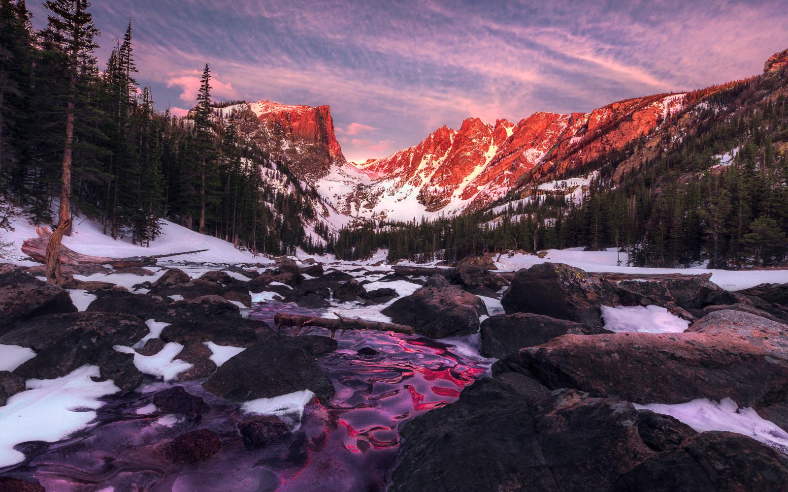 Dream Lake is truly a dream location to see and photograph. If you’ve never been, just go … However, if you plan on taking in the scene during the winter/snow months, understand your ‘dream' will include many layers of clothing and temperatures and windchill that can be in single digits with 20-30 MPH winds coming down the mountains and into your face … Those where my ‘dream' conditions during this gorgeous and chilly alpine glow sunrise.
I was the first one at the area on this morning, as my buddy and I arrived when the stars where still dominating the sky overhead. And before snapping any of those nighttime beauties, I made sure to set up my location for sunrise, to ensure I had the exact location within the boulders that I wanted. After the blue hour began and the stars were disappearing and more people started arriving to the area, I moved to my spot on the ice and hunkered down on the frozen water for a while. And since I wanted the lower angle with the ice and boulders as my foreground, helping lead you into the scene and then back to the mountains in the distance, I needed to basically lay on the ice during the entire time. Would I have changed anything on this morning? Heck no, because when the pink alpine glow began its show on the mountains, the cold seamed to just disappear.