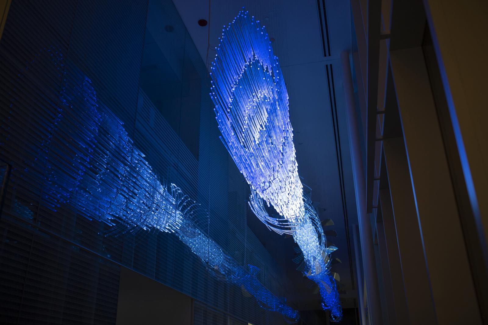 Nighttime view, illuminated.
Permanent installation at the Fascitelli Center for Advanced Engineering, College of Engineering, University of Rhode Island, Kingston, Rhode Island
Commissioned by the Rhode Island State Council on the Arts and University of Rhode Island
Light Pressure is a visual narrative that conflates mechanical, electrical, and optical phenomena, specifically blending pressure and fluid dynamics with the visuality of collimated light and force fields. Traveling down the corridor, a gestural vortex gives way to a linear flow of light encircled with curving glass and planar sheets, referencing magnetic fields, turbine blades, and supercavitation.
Light Pressure is created with suspended glass rods and curved canes. The rhythmic layers of glass create varying areas of density and sparsity to form an energetic, gently curving beam as the lens-like ends catch and bend light. Light Pressure is a site specific work, appearing differently depending on the audience’s viewpoint and time of day. Hand made from clear glass with embedded mica flakes to reflect and refract light, Light Pressure is semi-transparent and sparkling. In this way, Light Pressure is a shifting spectacle, while also receding into the architecture to become almost invisible.
At night the sculpture comes alive with dynamic custom lighting. Local tide and temperature data forms a basis of the machine-learning program, creating pulsing, sweeping, and spiraling patterns of shifting chroma and luminosity across the 1600 dimensionally mapped LEDs. This light is reflected off the interior glass wall and exterior curtain wall, creating an immersive feeling of deep waves and sparkling current as light pulses travel along the length of the sculpture.