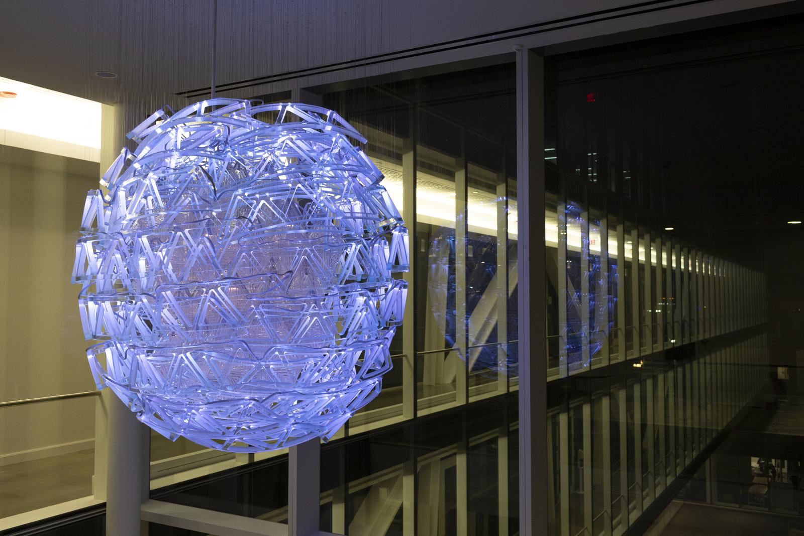 Night Image, Illuminated.
West Lounge. Fascitelli Center for Advanced Engineering, College of Engineering, University of Rhode Island, Kingston, Rhode Island
Commissioned by the Rhode Island State Council on the Arts and University of Rhode Island
Our site-specific sculptures, Droplets, investigate the elaborate forms and mass of nanoparticles while considering the atom and quantum mechanics as the driving force in our technology driven world. Droplet will serve both as an inspiration to those in the building, and as a beacon to campus, bridging the arts and humanities with engineering.
This work is made of both mold blown and kiln formed glass. The overall shape is a compressed toroid to maximize the double height ceiling in the lounges. The center of the sphere is made of mold blown lensed diamond shapes, while the outermost rings are made of waterjet cut and kiln formed glass trusses. In both the blown and kiln formed objects, we designed and made custom molds with the aid of 3D printing and traditional processes. The form and techniques used relate to the material engineering of nanostructures, where traditional truss structures are employed in increasingly complex ways.
The visible texture of the glass reveals the ways in which they were made, giving viewers access to the making process, while also providing a surface to refract light. During the day, the glass is translucent, catching light, while at night, the sculpture comes alive with dynamic light inspired by quantum entanglement and electron spin. With each piece of glass being its own light node, the trusses will seem as if they are spinning around the core of diamond lanterns. The three Droplets in the lounges and Bliss Hall being are connected together through the void of space, with the multithreaded lighting program creating visual dialog among the three pieces.