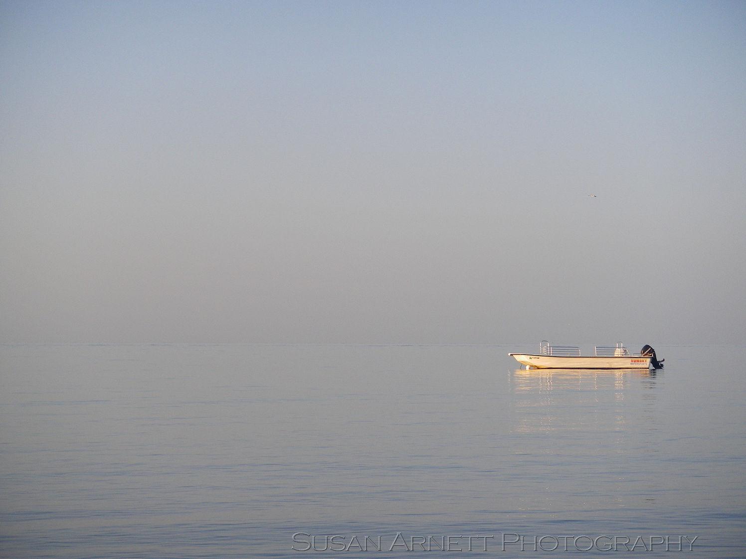 The empty sky and glass-still ocean melt together behind a small, lone, unoccupied boat as the sun rises, sending out golden rays, exuding a feeling of calm and surealness.