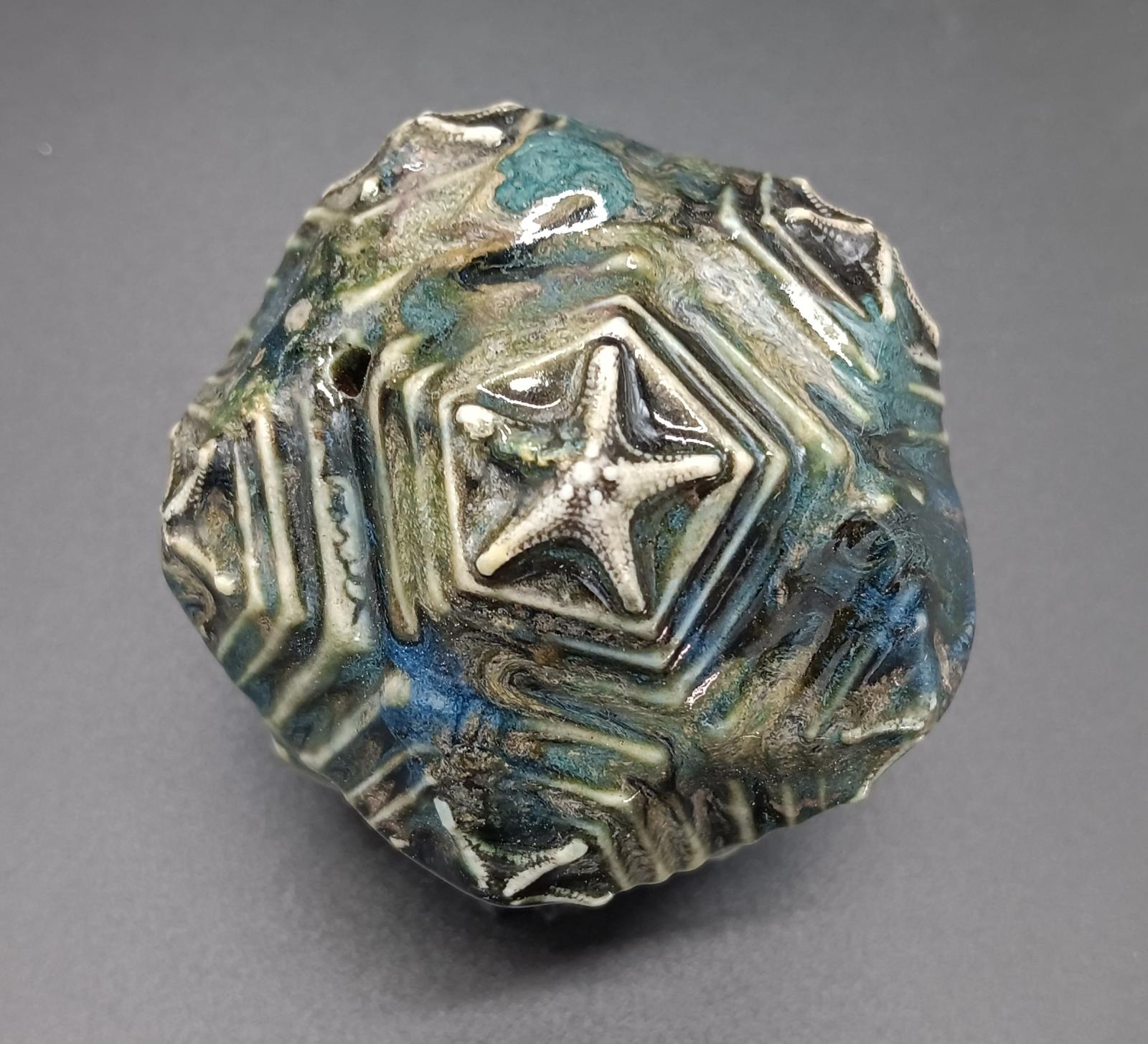 A porcelain dodecahedron, each of the 12 facets stepped upwards to a starfish expression. This is a fabrication, taking a full day to make. It is a reference in part to radiolarians, microscopic creatures that live is salt water, and in part to mineral growths, or perhaps biological processes that give rise to patterns. Starfish are stained black, with many layered stoneware glazes in green and blue to reference to sea. Fine example of a rare item that is one of a kind.