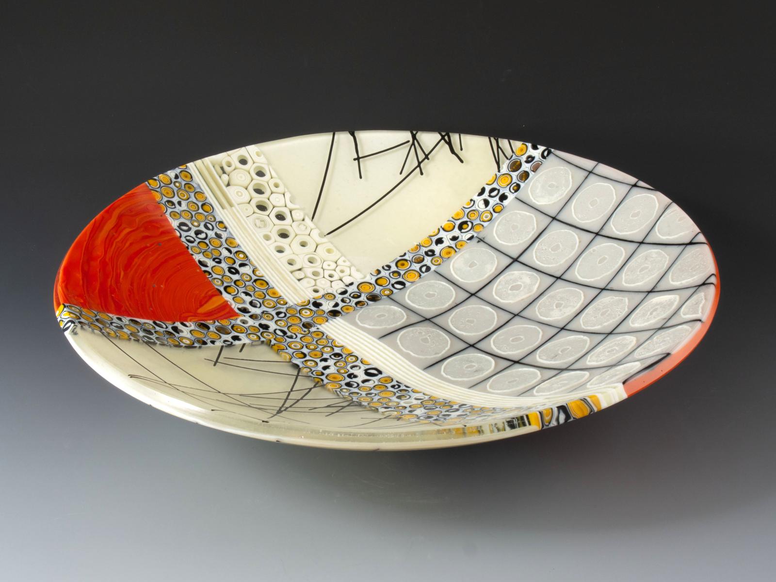 Bright red-orange ripples, sparkling iridescence, and complex patterned murrine abut rings of glass powder atop a grid of gray tiles seamed with black mortar. In contrast with the glossy smoothness of the rest of the bowl, the rings have irregular edges, thick rims, and stand off the surface to make the piece very tactile. You'll want to run your fingers over them every time you walk by.

The glass cylinders in this bowl are sections individually cut from hand-pulled glass cane, which I create by heating glass to 1500 degrees and pulling it into rods while molten in a modern-day adaptation of the 16th century techniques of the Murano glassmakers.

Fused, cold worked, and slumped.