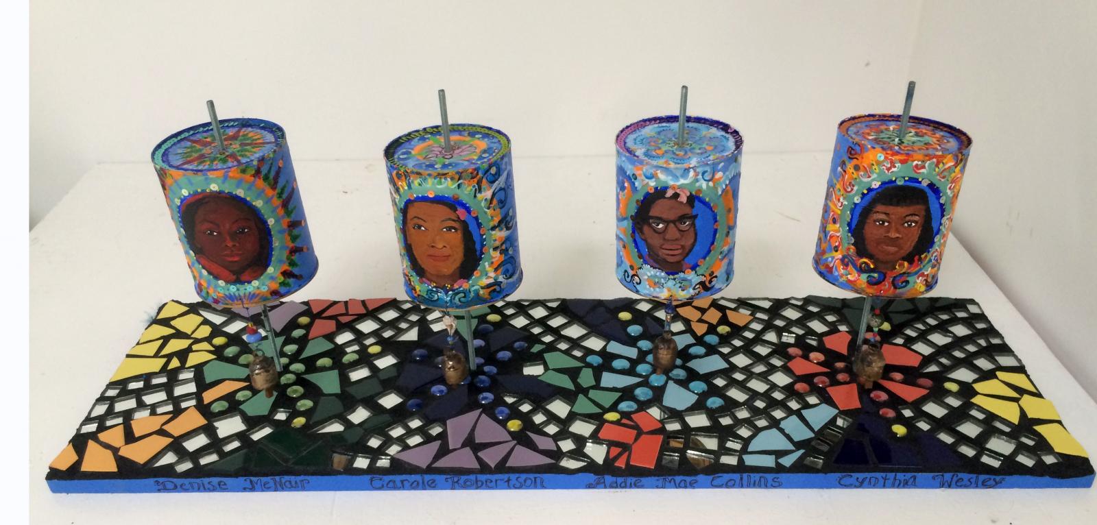 Please turn these Buddhist inspired prayer wheels to focus or meditate on compassion and healing. Turning the wheels clockwise creates energy by which to eliminate destructive energy.

The four little girls painted on the wheels are Addie Mae Collins, Cynthia Wesley, Carole Robertson and Carol Denise McNair who perished in racist terrorist bombing of The 16th Street Birmingham Church in 1963. While working on this piece, another act of racist terrorism occurred, the Charleston Church Shooting in 2015. In response, I added the names of the eleven victims.
There is no more room to add the ever expanding roster of names and faces taken from this earth through violence rooted in racism.

Please turn the wheel to counter the political spoken and unspoken effects of hatred and racism.
The mantra, Om Mani Padme Om, (Lotus Mantra) is traditionally repeated.

Just as a lotus grows forth and pushes out from the mud,
may I emerge out of the current culture of despair and violence. May I rise into beauty and compassion.

May I act on this.
Rise up.
