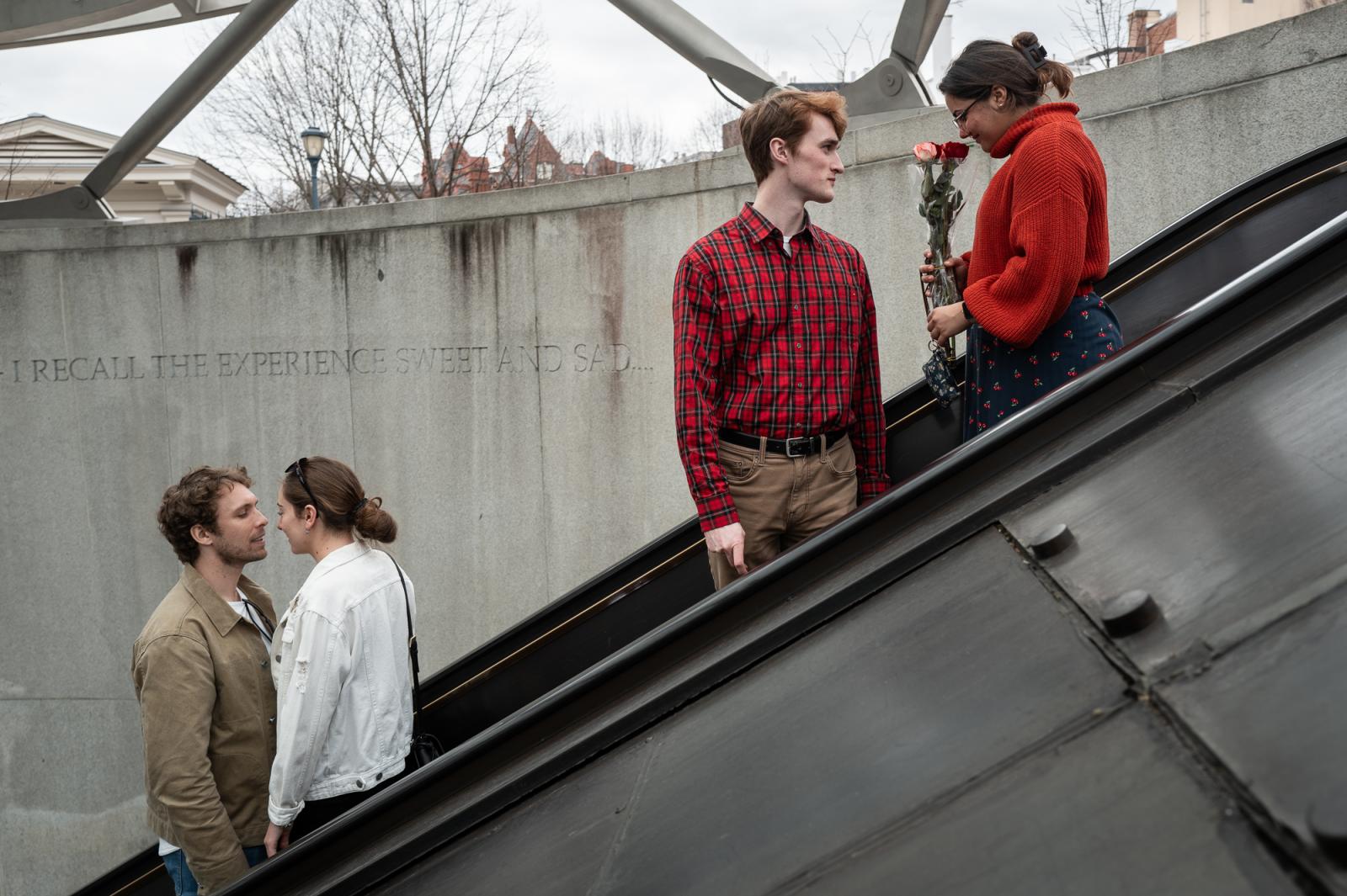 Four people, two groups of two facing each other, descend into the Washington, D.C. Metro at the Dupont Circle station. On the right, a woman in a red sweater holds long stemmed roses and looks into them while a young man, also dressed in red looks at her.  On the left, a young man with a beard and wearing a tan jacket faces a young woman with her hair pulled back and standing on the step directly above him. Behind them on the circular stone wall of the entrance is a portion of a quote by Walt Whitman: "I recall the experience sweet and sad ..."