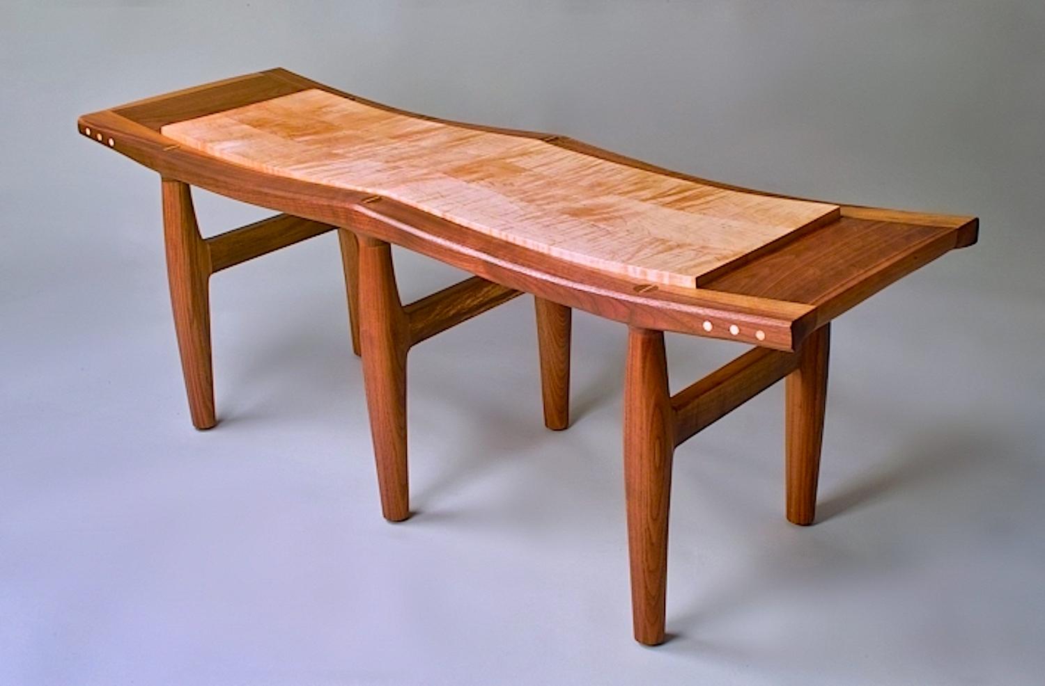 Straight grained black walnut bench, supporting a floating top made of a shimmery curly maple that seems to ripple in the light.  Exposed joinery and maple pegs on the frame accent the piece.