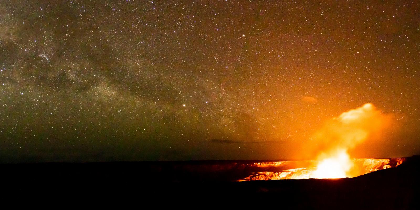 An erupting Kilauea Volcano sends smoke acsoss a star-strewn sky, highlighted by the Milky Way.