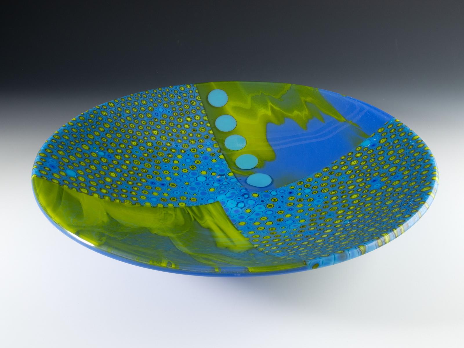 Land and sea meet and mingle, bright springy greens swirling with oceanic blue. Thin discs of paler blue float above, casting a shadow onto the sea floor. Like a drift of anemones, a gathering of cylinders of hand-pulled cane divides the watery quadrants.

The glass cylinders in this bowl are sections individually cut from hand-pulled glass cane, which I create by heating glass to 1500 degrees and pulling it into rods while molten in a modern-day adaptation of the 16th century techniques of the Murano glassmakers.

Hundreds of them are placed by hand onto a ceramic surface, standing together in a tight formation, and heated until they soften and puddle together to form a slab.

Fused, cold worked and slumped.