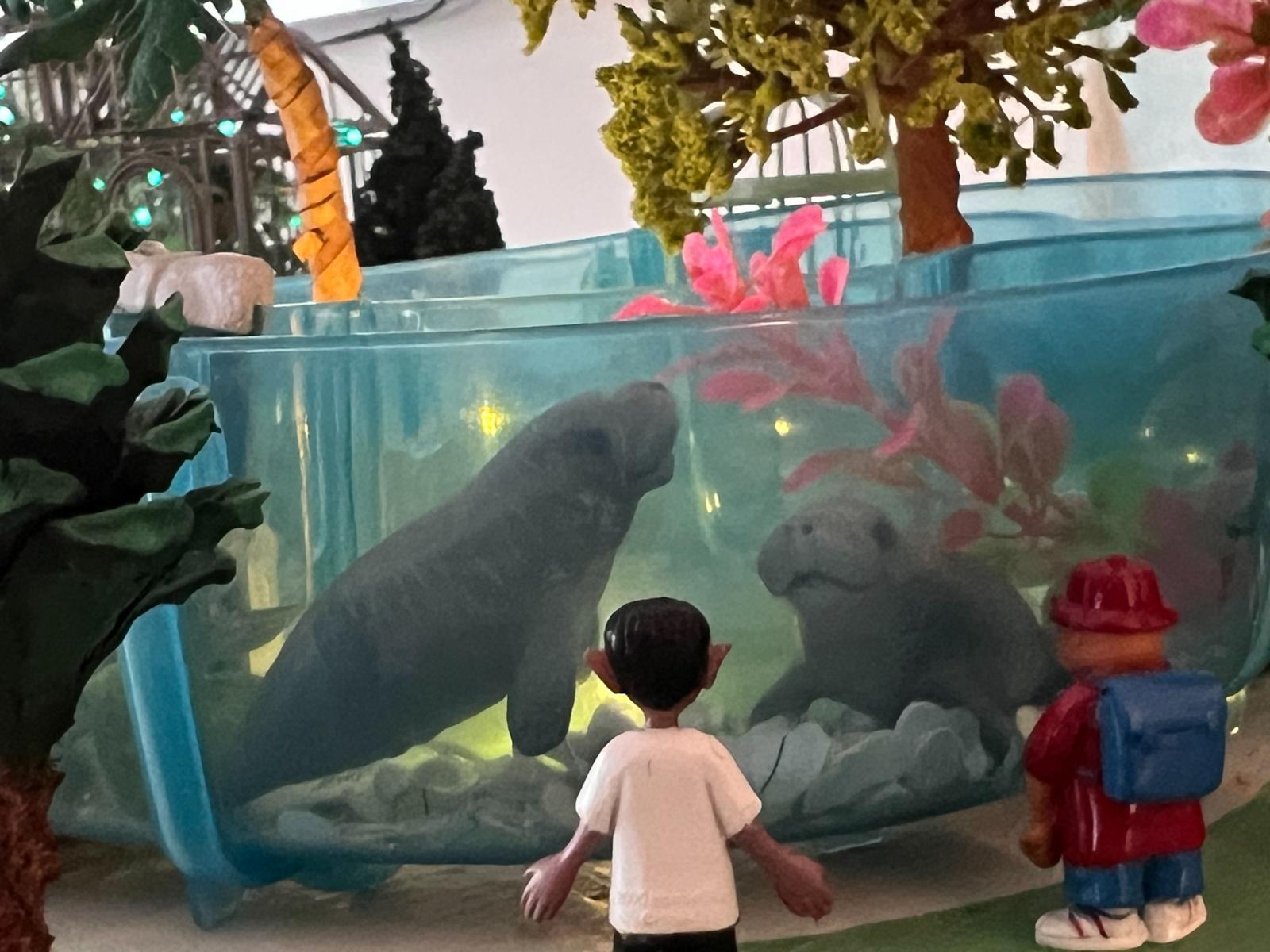 This manatee exhibit was meticulously crafted using a variety of repurposed materials, transforming discarded items into a vibrant and imaginative habitat. The base of the exhibit was formed from a discarded child's water toy. Hole punches were utilized to create texture and detail.

Flora in the exhibit includes discarded aquarium plants, complemented by discarded plastic flowers that were reshaped and given new life with spackle. The trees within the exhibit were painted with acrylics, and their trunks were fashioned from discarded packing paper. The unique fronds for the palm trees were ingeniously created from mandarin orange peels.

Further natural elements include a pine cone and sumac buds, integrated to enhance the exhibit's organic feel. Plastic animals and miniature plastic people from gumball machines populate the scene, adding scale and narrative to this environmentally conscious display.

