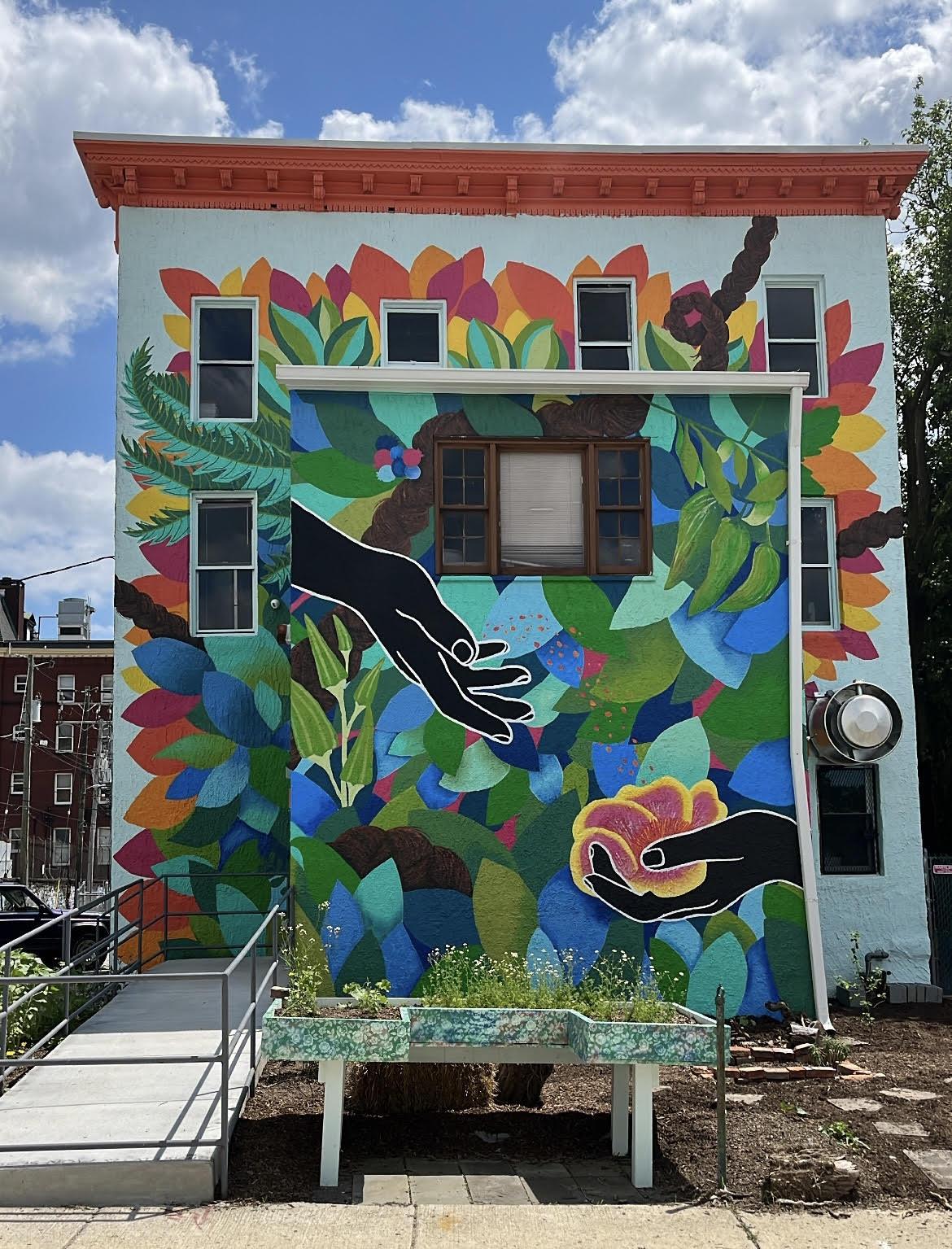 This mural follows the story of the seeds that have shaped Southern cuisine. The seeds were braided into enslaved people's hair. In the center, Black hands share those seeds through the generations. The seeds land into a giant glowing Mimosa blossom. A braid weaves through the entire piece. Okra stands proud in the front with Baltimore native a fish Peppers hanging from the corner. Basil, cannabis, and plantain broadleaf leaves peek out at the top. The mural stands in a garden of healing and edible plants planted by chefs Cat and Kiah at Our Time Kitchen with the community. Neighbors helped to paint in the bottom layer of leaves on a painting day facilitated by the artist. This project was made possible through the New Public Art across Maryland grant funded by MSAC.
Location: 117 W 24th St. Baltimore, MD