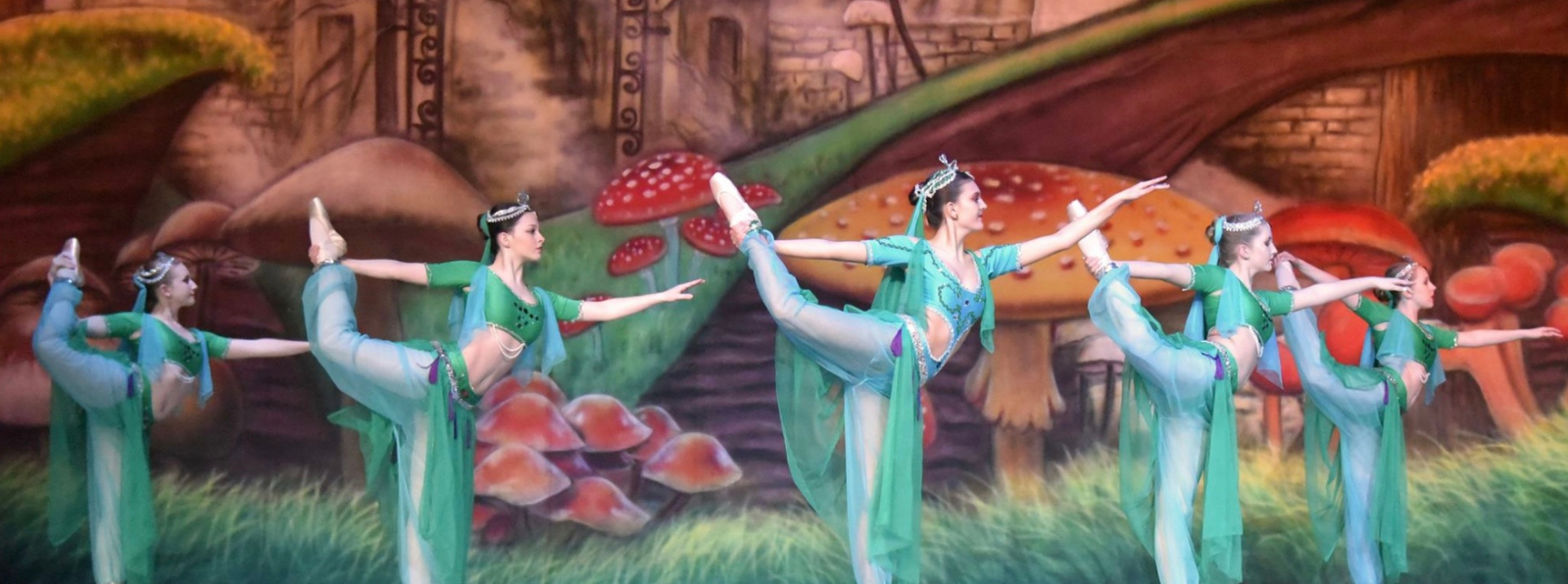 Five costumed people lift their right legs up while performing in front of a painted scene.